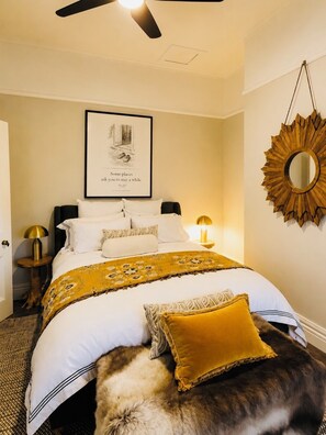 A bedroom with a bed, bedside lamps, a framed quote on the wall, and a sunburst mirror.