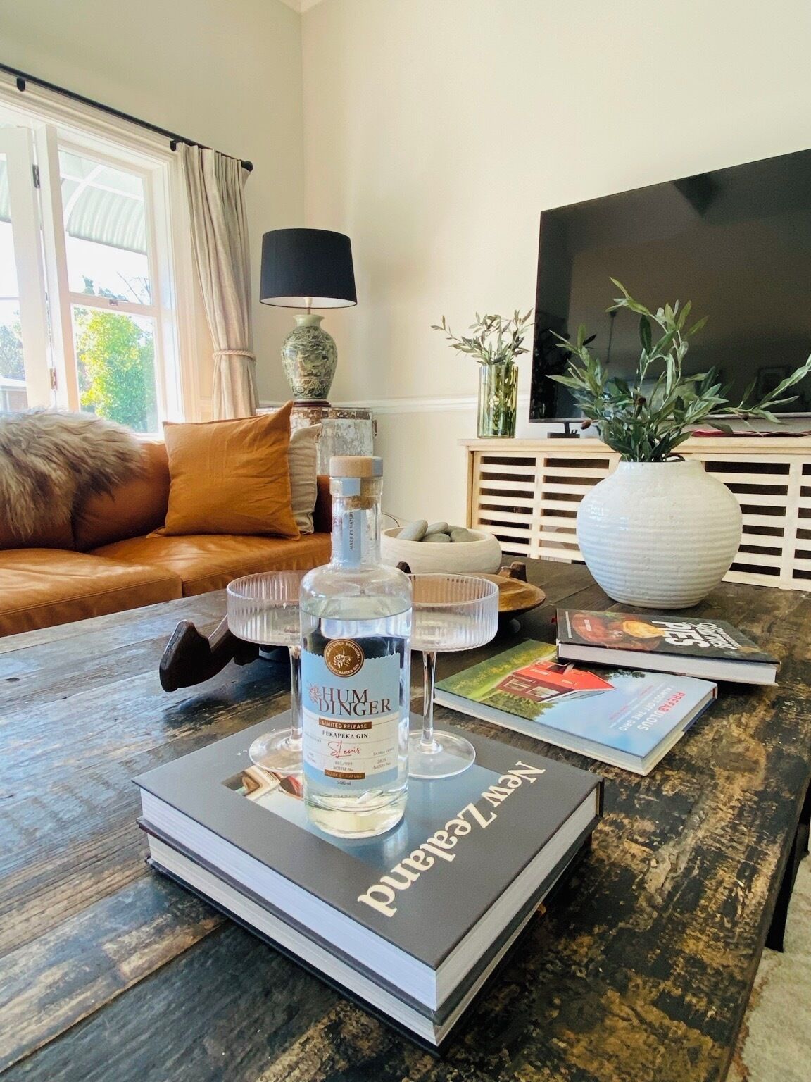 A living room with a sofa, a coffee table with books and a bottle of gin, a TV, and a window with curtains.