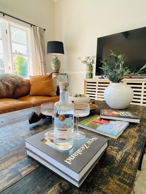 A living room with a sofa, a coffee table with books and a bottle of gin, a TV, and a window with curtains.