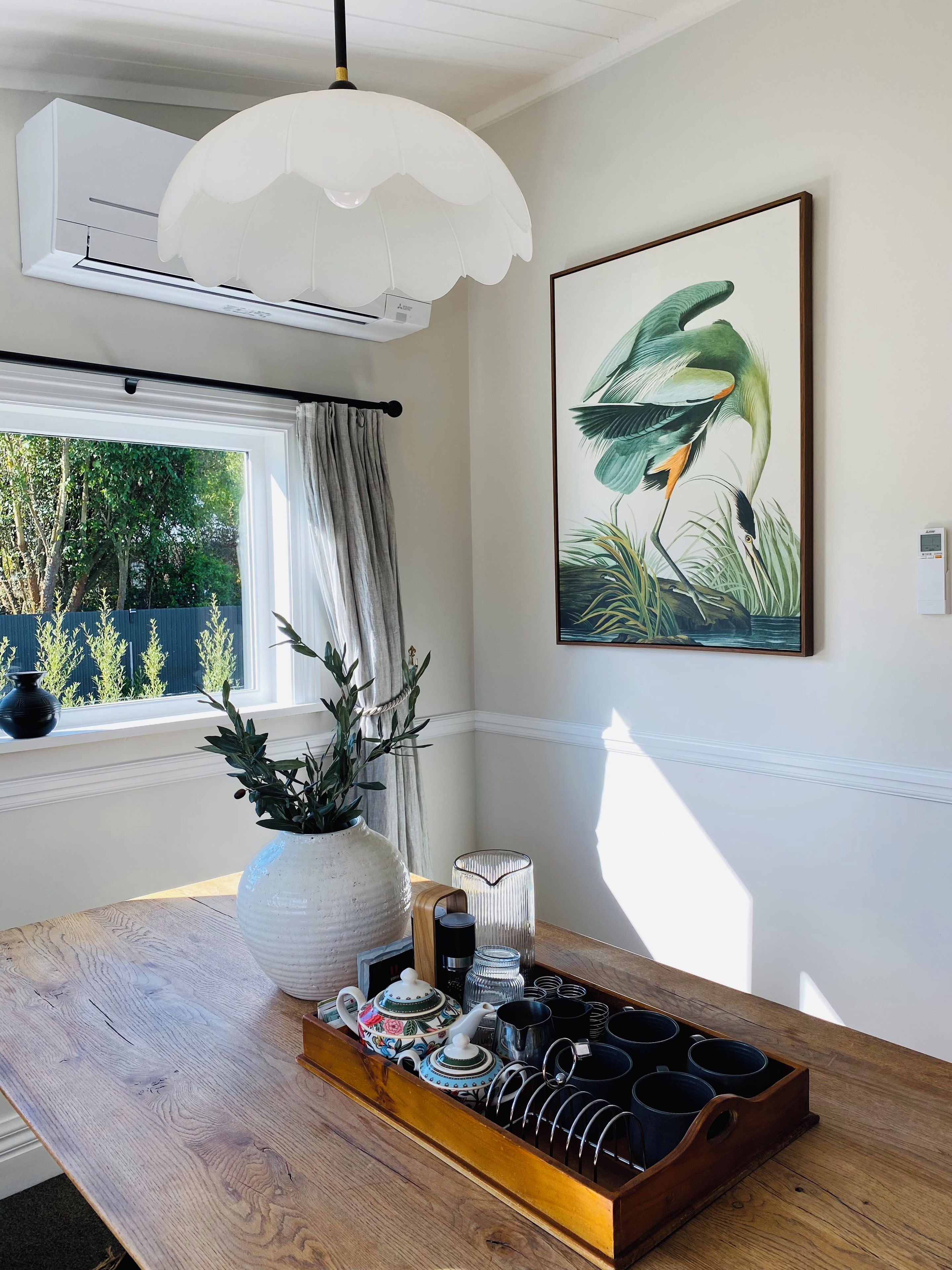A room with a wooden table, a white vase with green plants, a framed bird painting, and a window with curtains.