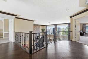 A spacious interior with a wooden floor, a staircase with a metal railing, and a view of the outdoors through large windows. - Luxury Aspen Woods Rental | 5 Bedrooms | 4.5 Bathrooms | 5,500+ SF (Calgary)