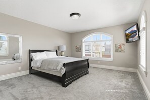 Ein Schlafzimmer mit einem Bett, einem Fenster mit Jalousien, einem an der Wand montierten Fernseher und einer Deckenleuchte.