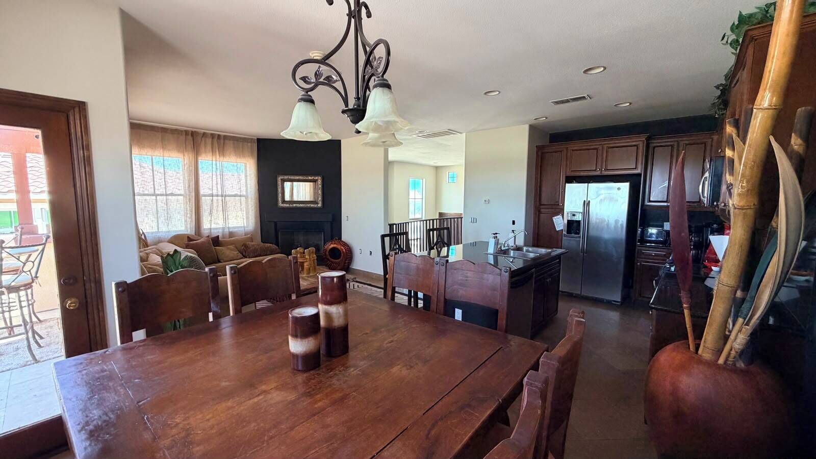 Uma área de refeições com uma mesa de madeira, cadeiras e um candelabro. Dá para ver uma cozinha com geladeira e armários ao fundo.