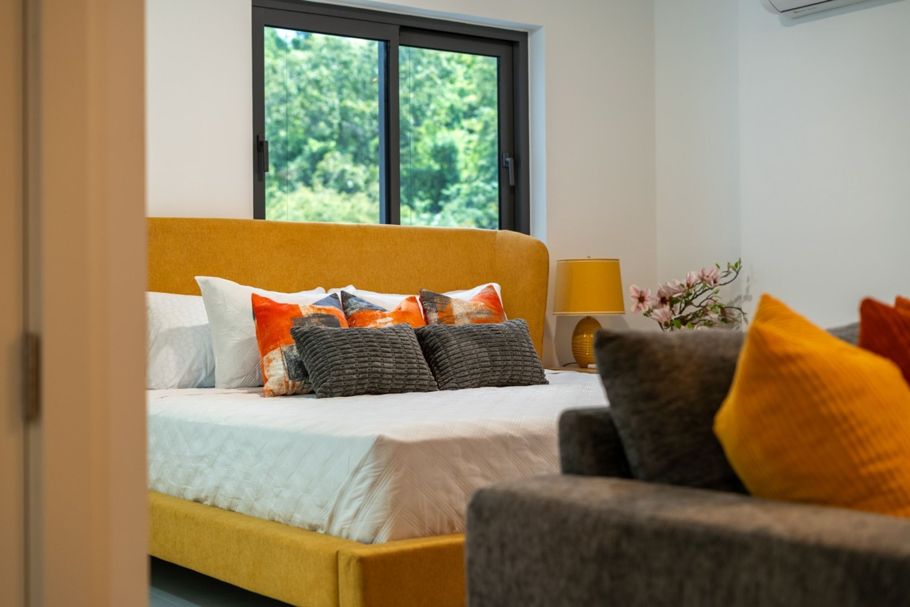 A bedroom with a bed, a nightstand, a lamp, and a window with a view of greenery.