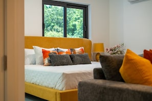 A bedroom with a bed, a nightstand, a lamp, and a window with a view of greenery. - SOL Euphoria Walk to Beach Town (Ocho Rios)