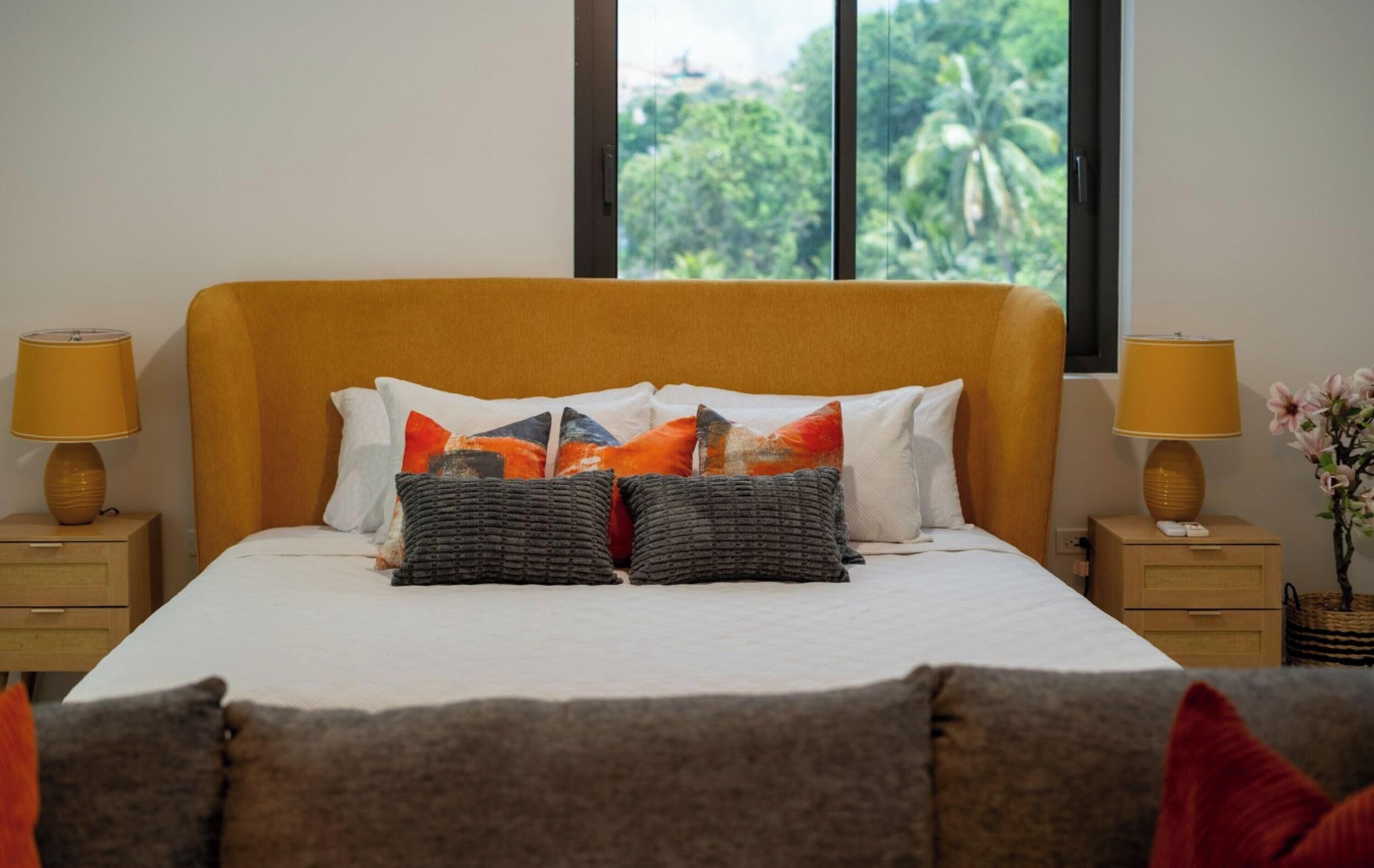 A bed with a white mattress, a yellow headboard, and multiple pillows in various colors. There are two bedside tables with lamps and a vase with flowers.