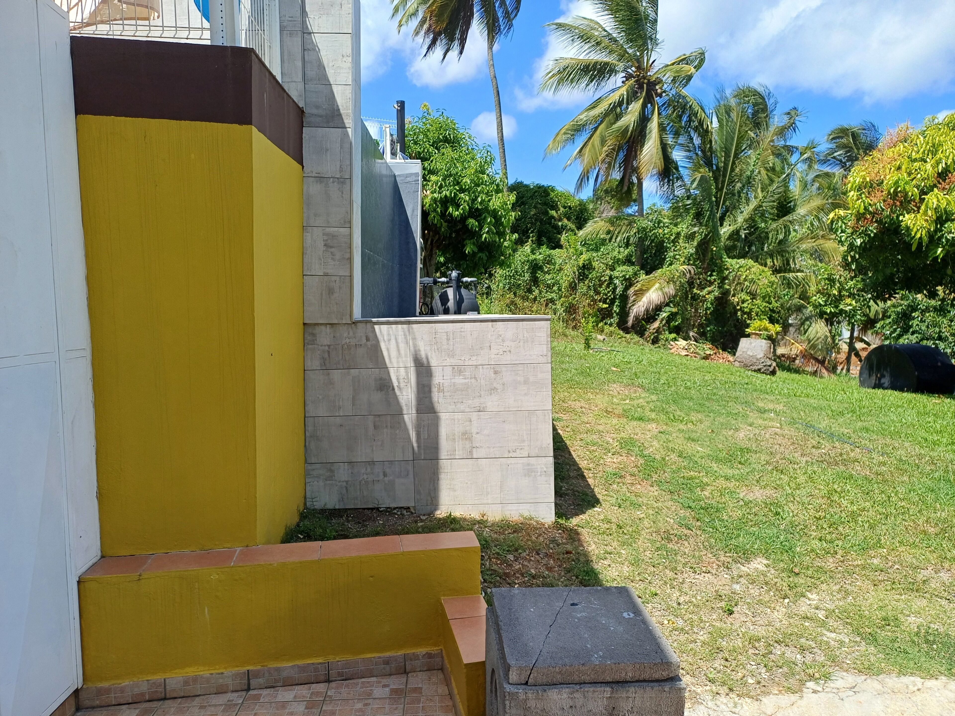 Un edificio con fachada amarilla y gris, un banco de hormigón y una zona ajardinada con palmeras.