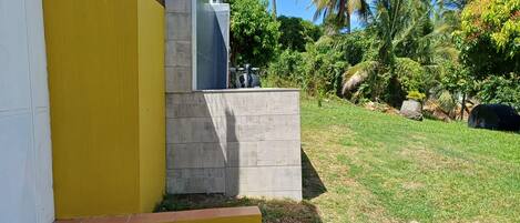 Un edificio con fachada amarilla y gris, un banco de hormigón y una zona ajardinada con palmeras.