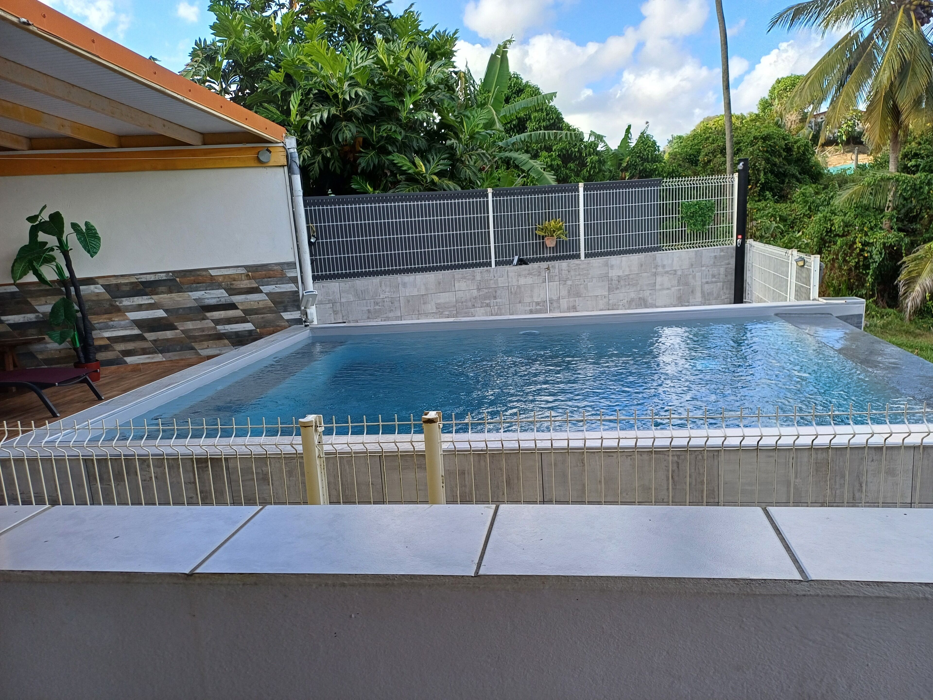 An outdoor swimming pool with a tiled deck, surrounded by a fence and lush greenery.