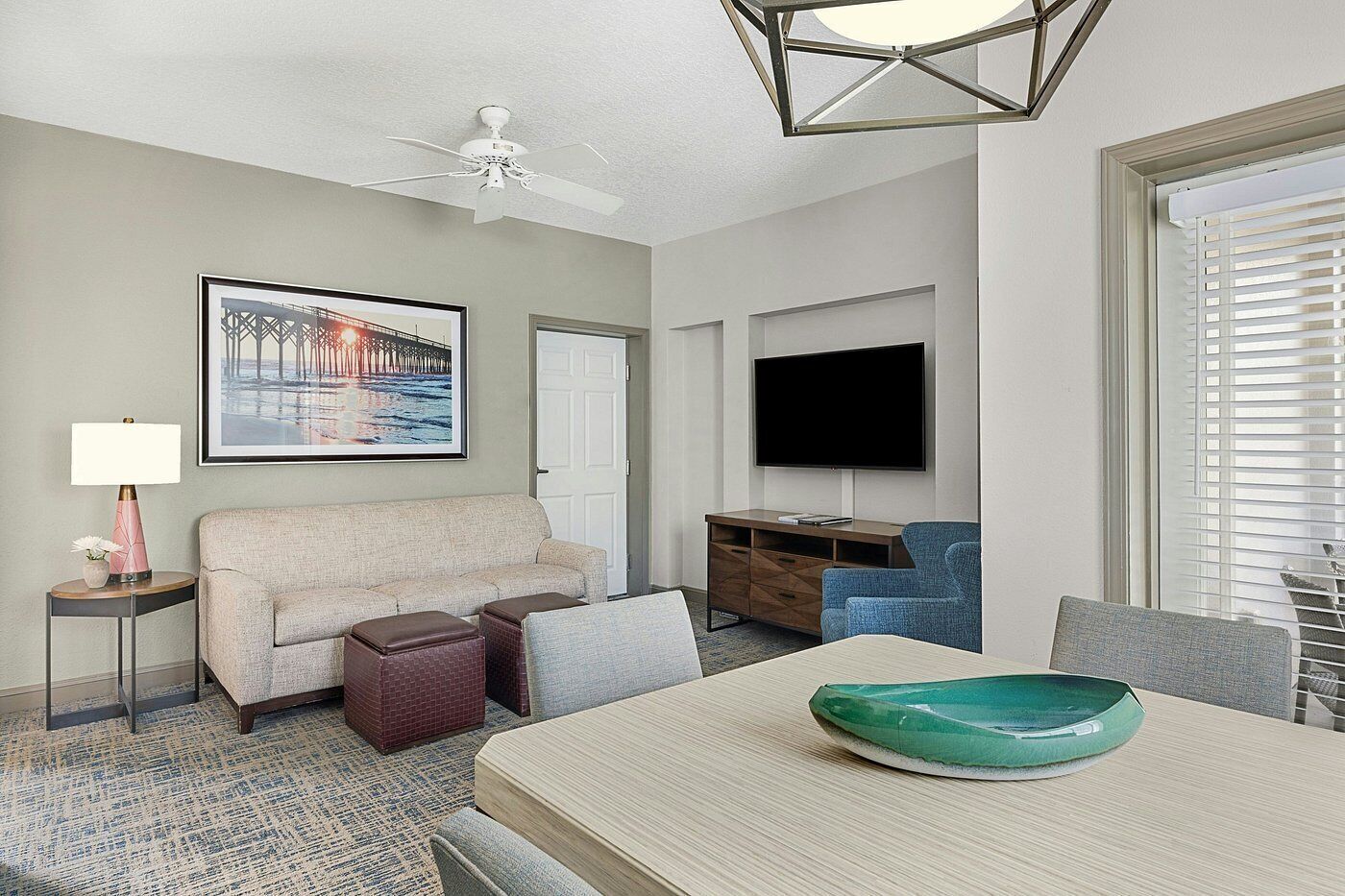 Habitación de hotel con sofá, una mesa de comedor con un tazón verde, televisor de pantalla plana, ventilador de techo y un cuadro en la pared.