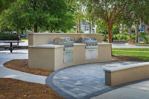 An outdoor barbecue area with a built-in grill and a cabinet, surrounded by a landscaped garden and a paved walkway. - Myrtle Beach | Group Beach Retreat | Pool + Hot Tub | 2BR (Myrtle Beach)
