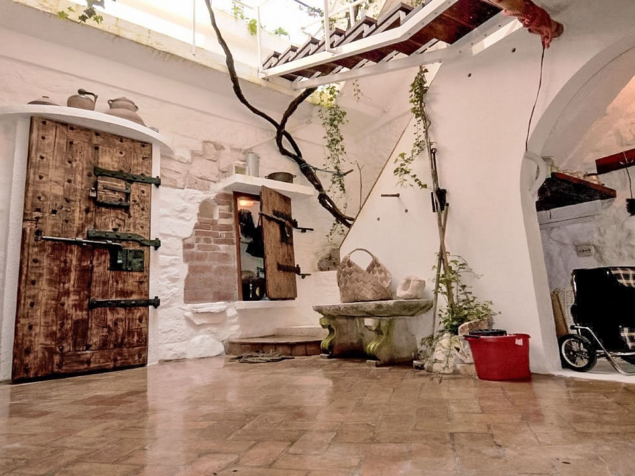 A courtyard with a wooden door, a stone wall, a hanging plant, and a red bucket.