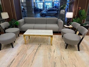 A modern lobby with a grey sofa, matching armchairs, a marble coffee table, and a glass wall reflecting the outside. - Sky View Hotel (Madinah)