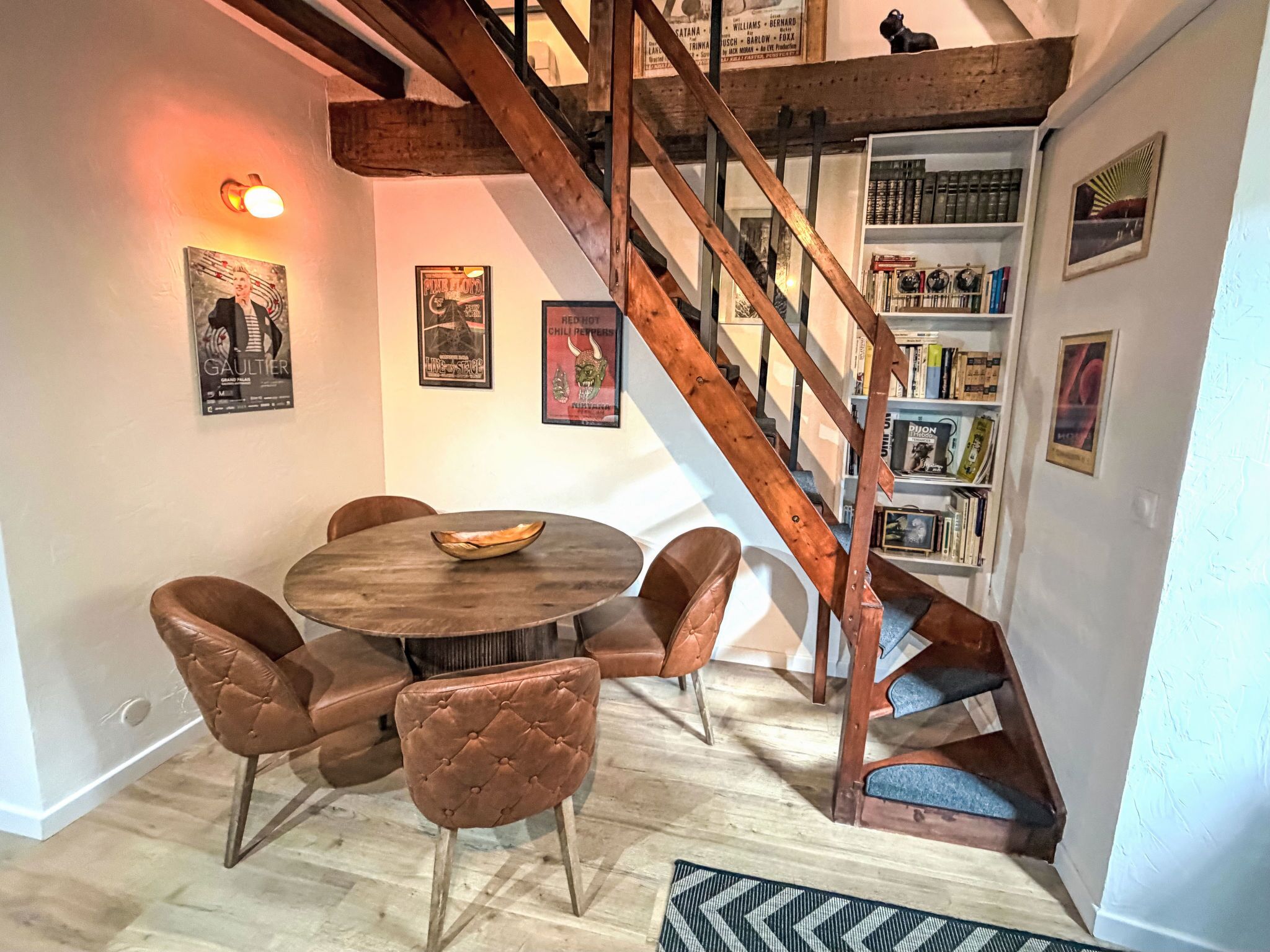Een eetgedeelte met een houten tafel en stoelen, een trap met zichtbare balken en een boekenplank vol boeken.