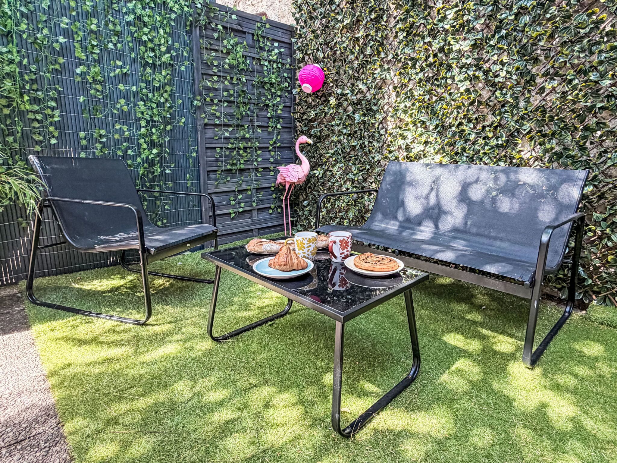 Een terras met een zwarte metalen bank en stoelen, een tafeltje met borden eten, en een decoratieve flamingo.
