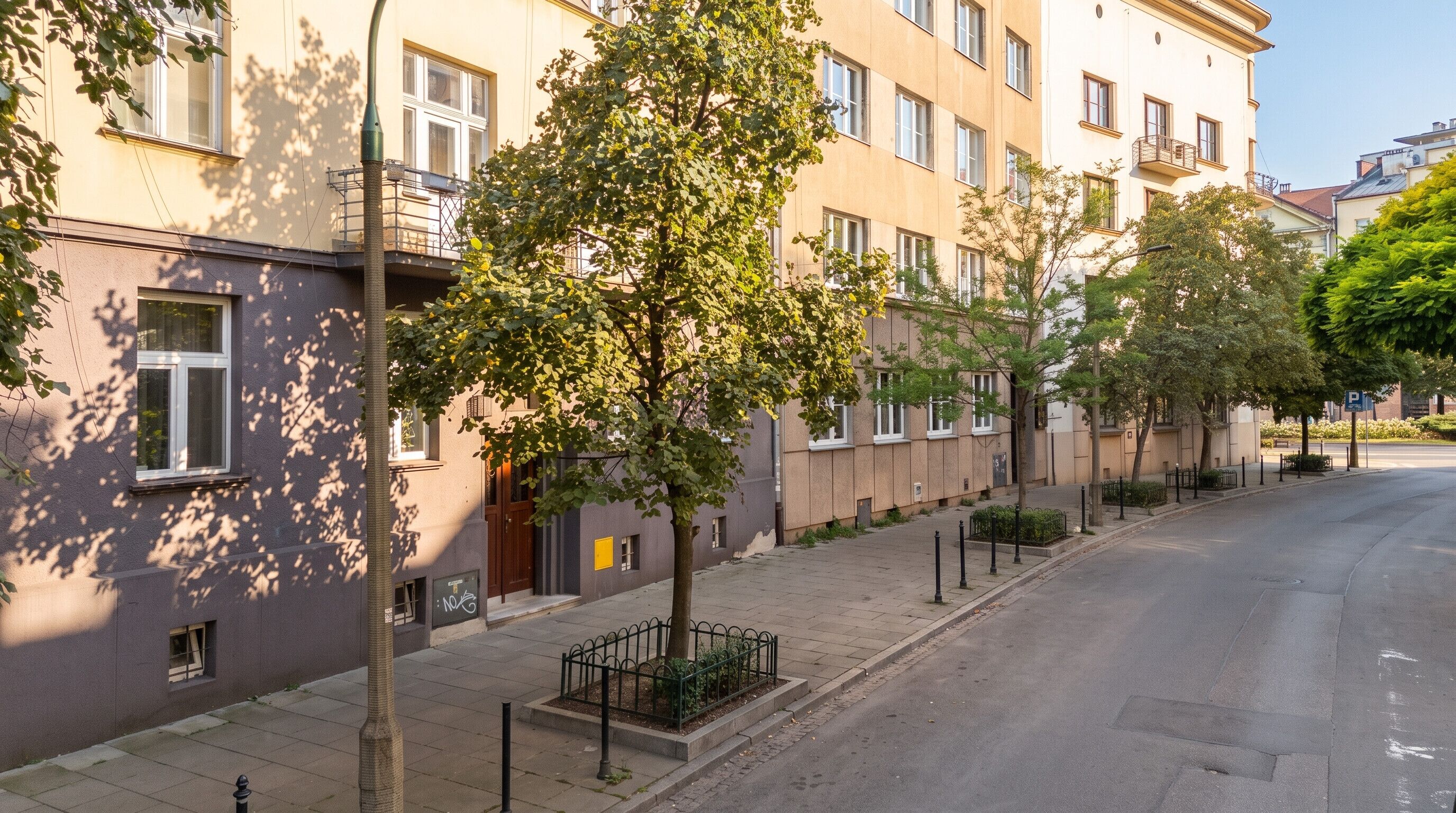 Eine Reihe von Wohngebäuden mit Balkonen, ein klarer Himmel und eine von Bäumen gesäumte Straße.