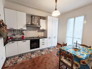 Eine kompakte Küche mit weißen Schränken, einem Gasherd und einer Spüle. Es gibt einen Essbereich mit einem Tisch, der mit einer sonnenblumenmusterigen Tischdecke bedeckt ist.