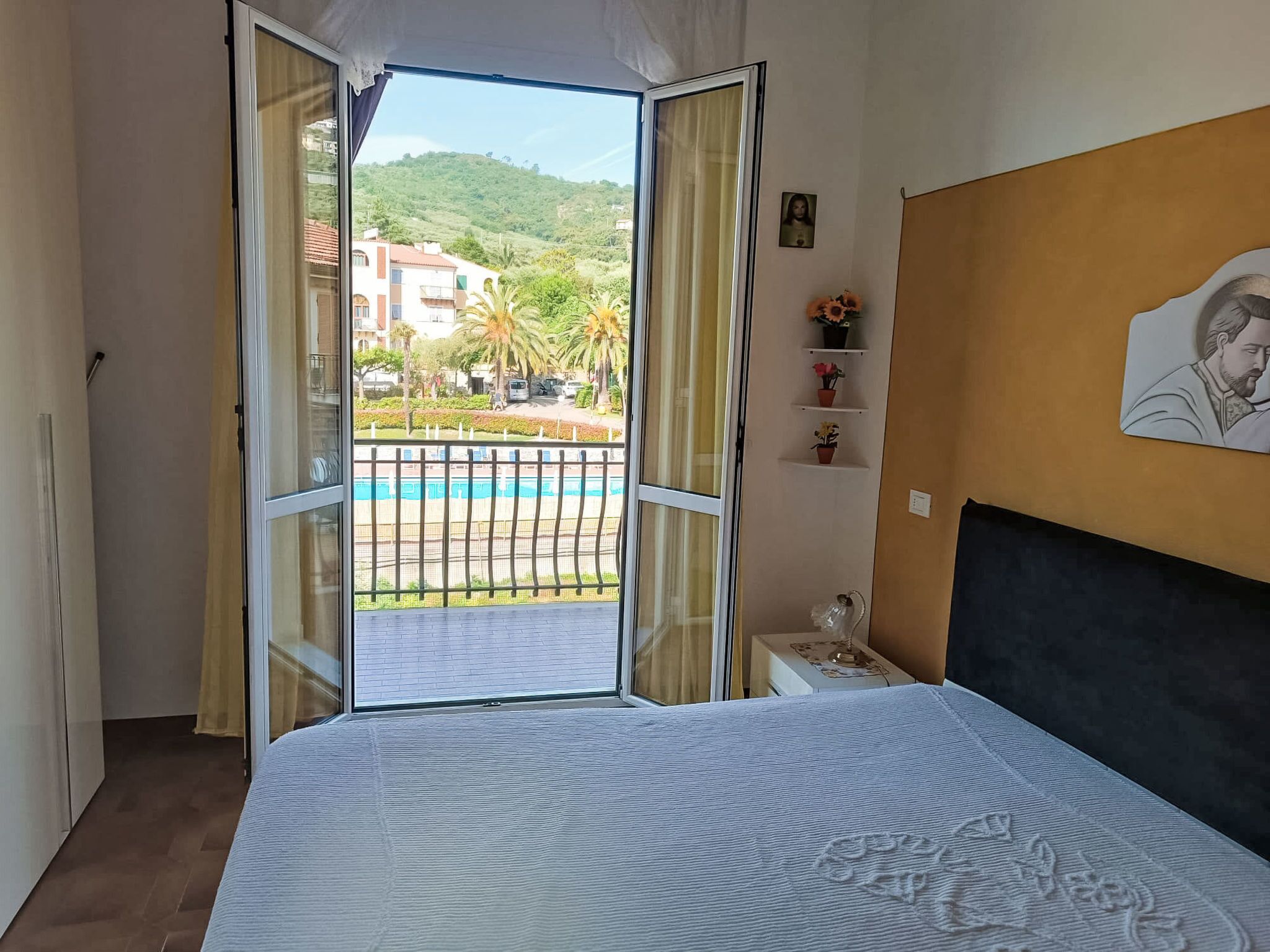 A bedroom with a bed, a nightstand, a painting, and a view of a pool and buildings.