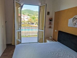 A bedroom with a bed, a nightstand, a painting, and a view of a pool and buildings. - Stella Marina by Interhome (Pietra Ligure)