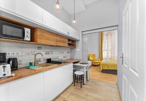 A modern kitchen with white cabinets, a microwave, and a sink. There is a dining area with a yellow chair and a bed with a yellow bedspread. - Topolowa Comfort Apartment (Kraków)