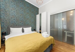 Quarto de hotel com cama, mesa de cabeceira, luminária, guarda-roupa e porta de correr para o banheiro.