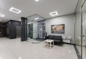 Un hall d’entrée moderne avec un ascenseur en verre, une table basse blanche, un canapé et une œuvre d’art représentant une carte géographique au mur.