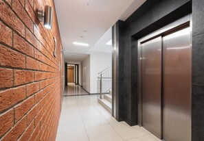 A hallway with a brick wall, a metal elevator, and a staircase with a metal railing. - Podgórze Boutique Studio (Kraków)