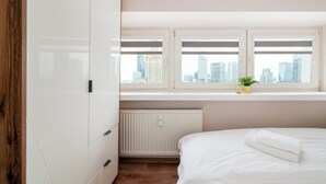 Quarto com cama, radiador, janela com vista para a cidade e guarda-roupa de madeira.