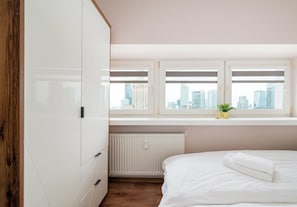 A bedroom with a bed, a radiator, a window with a city view, and a wooden wardrobe. - Zgoda 13 Modern City Apartment (Warszawa)