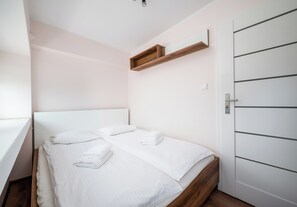 Un dormitorio con cama, dos almohadas, cabecera de madera, una puerta blanca y un pequeño estante sobre la cama.