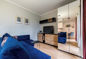 A modern living room with a blue sofa, a TV on a wooden cabinet, and a dining area visible through a glass partition. - Posnania Comfort Studio with Garden (Poznań)
