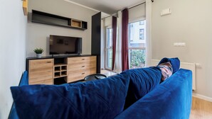 Ein modernes Wohnzimmer mit einer großen, blauen Couch, einem Flachbildfernseher auf einem Holzschrank und Blick auf ein Gebäude durch ein Fenster mit Vorhängen.