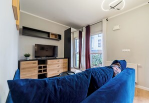 Ein modernes Wohnzimmer mit einer großen, blauen Couch, einem Flachbildfernseher auf einem Holzschrank und Blick auf ein Gebäude durch ein Fenster mit Vorhängen.