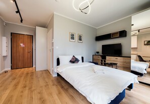 A modern hotel room with a wooden floor, a large bed with white bedding, a flat-screen TV, a wooden dresser, and a small table. - Posnania Comfort Studio with Garden (Poznań)