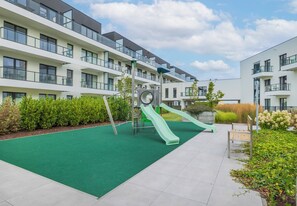 A playground with slides and a green artificial turf surface, surrounded by apartment buildings with balconies and large windows. - Zielonego Mostu Cozy Elegant Apartment (Szczecin)