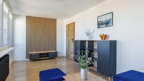 Una sala moderna con piso de madera, sofás azules, una mesa de centro de madera, un aparador negro con una planta y un frutero, y un cuadro de un paisaje urbano en la pared.