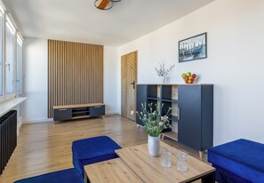 Ein modernes Wohnzimmer mit Holzboden, blauen Sofas, einem Holztisch, einer schwarzen Kommode mit Pflanze und Obstschale sowie einem Stadtbild an der Wand.