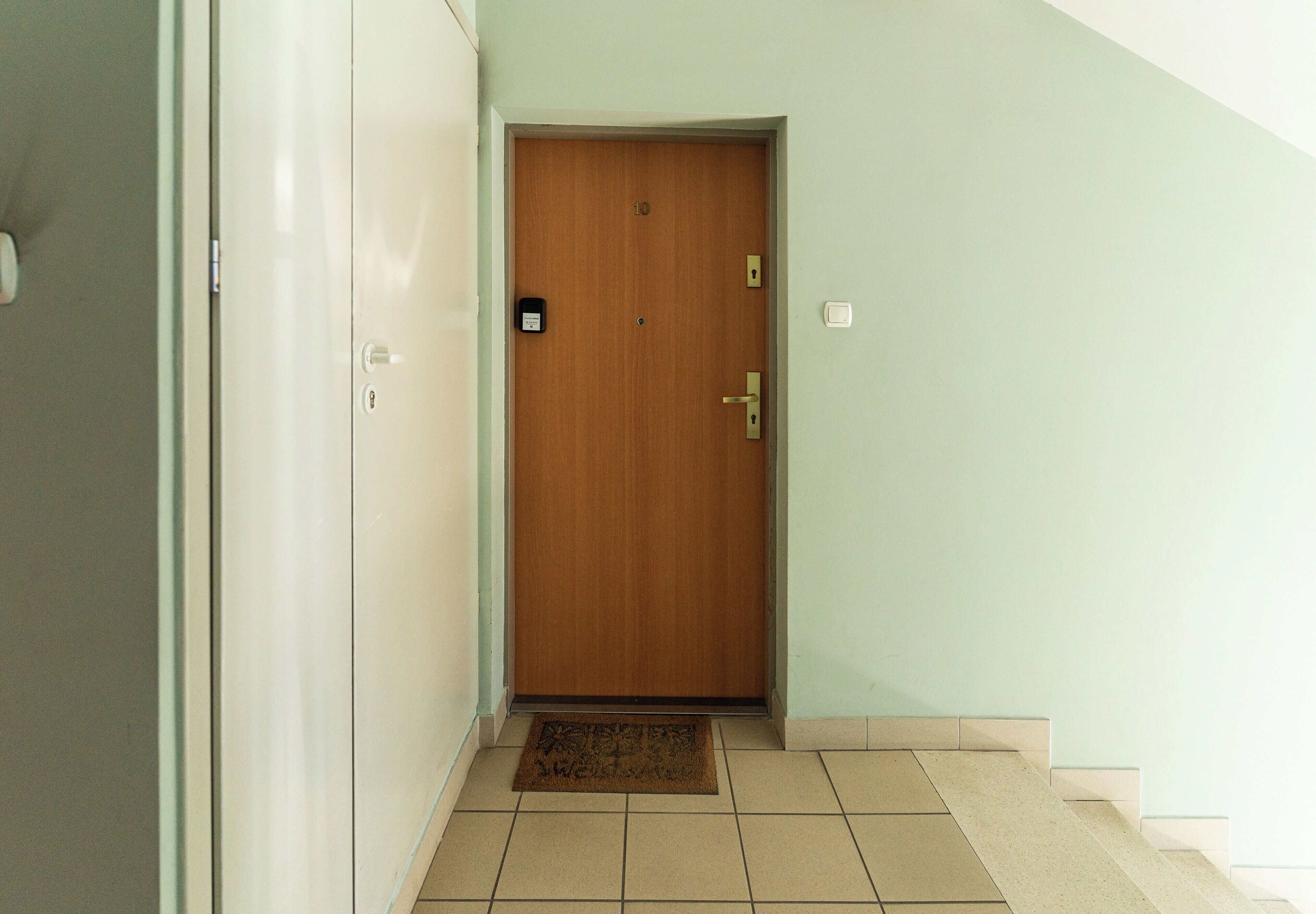 Un pasillo con una puerta de madera, un felpudo y un interruptor de luz en la pared.