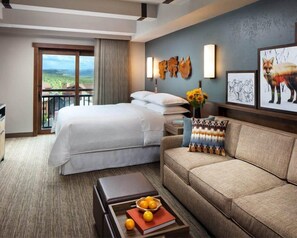 A hotel room with a large bed, a sofa, a coffee table with fruits and books, a balcony with a view, and wall art. - Steamboat Springs | Mountain Ski | Ski Access & Hot Tub (Steamboat Springs)