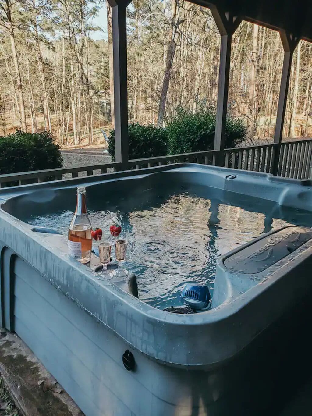 Un bain à remous avec une bouteille et des verres posés sur le bord, entouré d’une terrasse en bois et d’arbres.