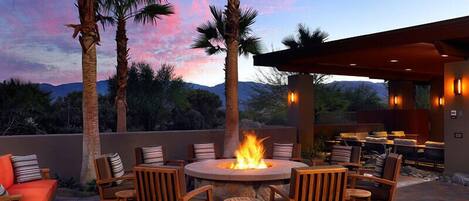 An outdoor patio with a fire pit surrounded by wooden chairs and tables, palm trees, and a view of the mountains.