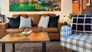 Un salón con un sofá de cuero marrón, un sillón escocés, una mesa de centro de madera y un cuadro grande de un molino de agua.