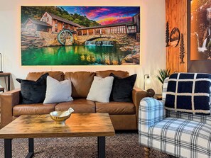 A living room with a brown leather sofa, a plaid armchair, a wooden coffee table, and a large painting of a watermill. - Creekside walk to downtown, landing, king bed (Branson)