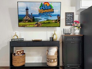 A television mounted on a wall displaying 'Aha Moments Property Management' with a scenic view. Below, a black console table holds a sign, a bowl, and baskets. A cabinet with a drawer is to the right. - Creekside walk to downtown, landing, king bed (Branson)