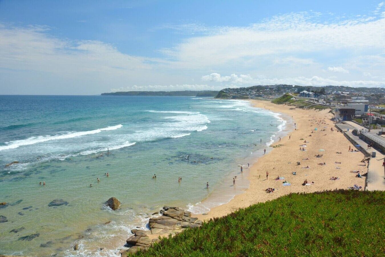A coastal beach with clear waters, a sandy shore, and a view of a town in the distance.