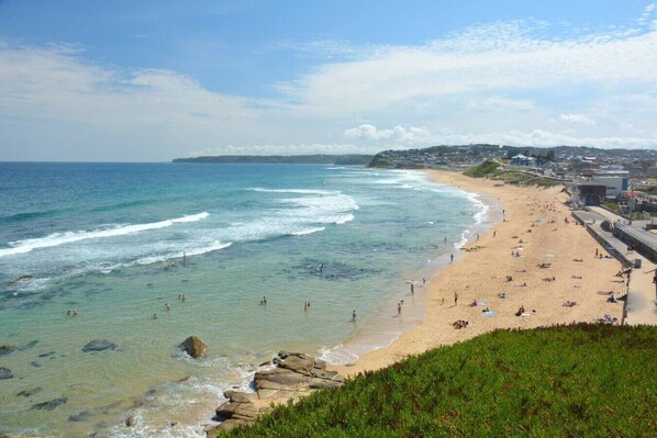 A coastal beach with clear waters, a sandy shore, and a view of a town in the distance.
