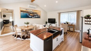 Eine moderne Küche mit Kochinsel, Essbereich mit rundem Tisch sowie ein Wohnbereich mit Sofa und Fernseher.