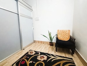 Una habitación con piso de madera, una silla negra con cojín estampado, una planta en maceta y un tapete decorativo.