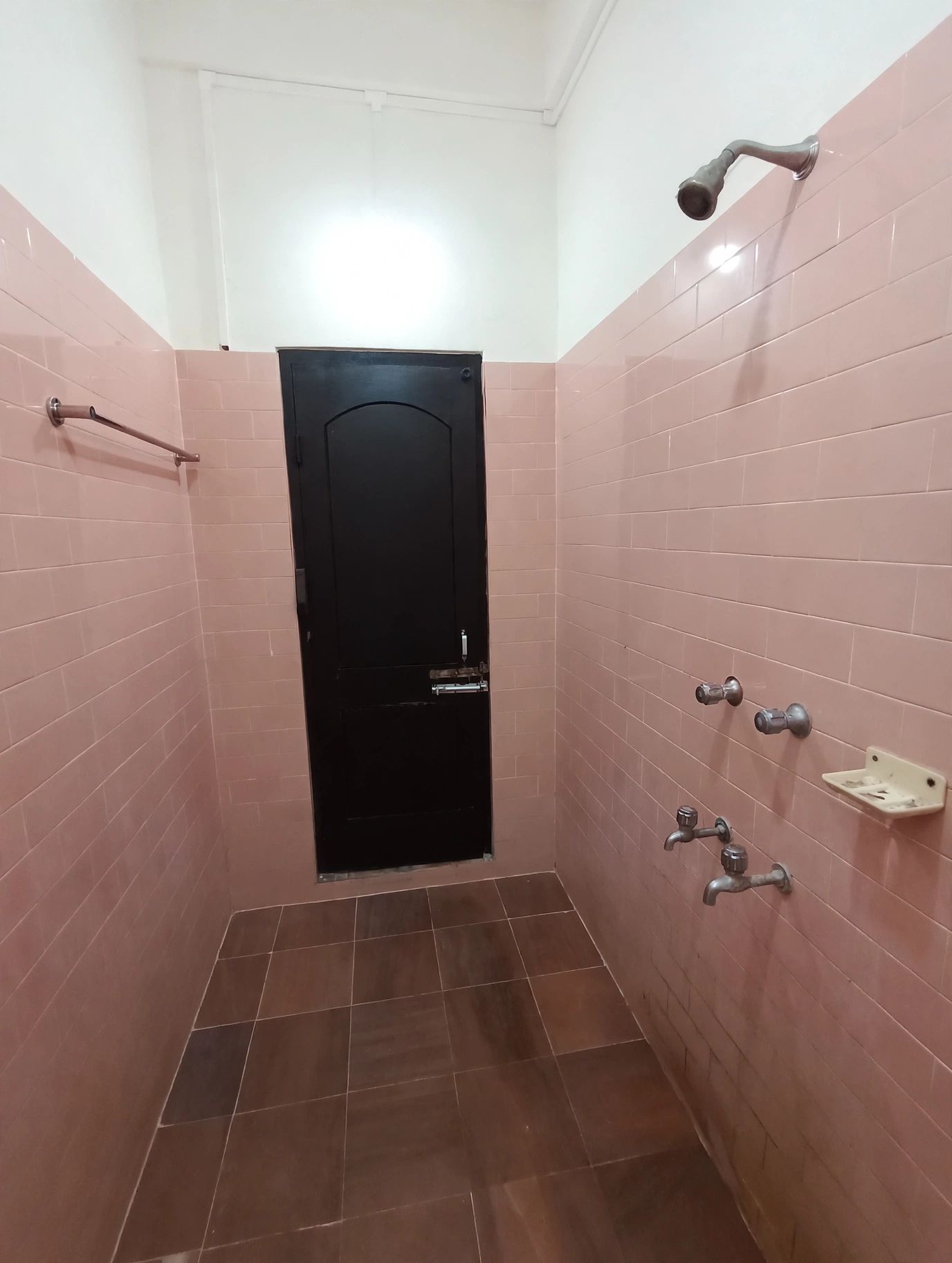 Une salle de bain avec des murs recouverts de carreaux roses, une porte noire et un espace douche équipé d’un pommeau de douche à main.