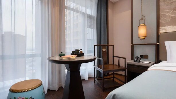 A hotel room with a bed, a desk with a telephone, a chair, a small table with books, and a window with sheer curtains.
